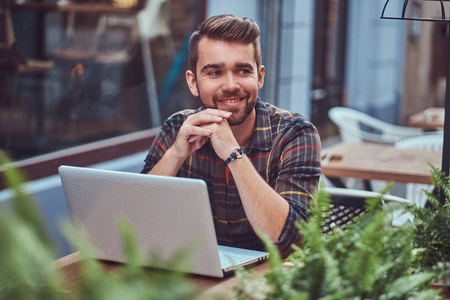 一位微笑的年轻自由职业者的肖像，发型时尚，胡须穿着羊毛衬衫，坐在户外咖啡馆里的笔记本电脑上工作。