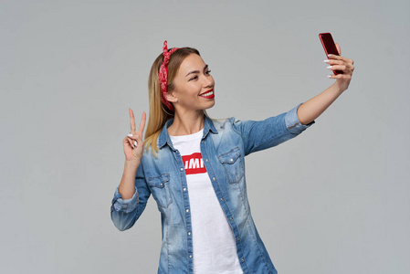 年轻的时髦女人采取自拍智能手机, 广泛的微笑和显示胜利和和平的标志