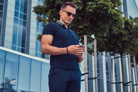 一个英俊的时尚男人，戴着黑色T恤和裤子的太阳镜，有一个时尚的发型，在现代城市的智能手机支架上对着摩天大楼读着一条信息。