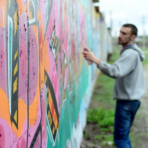 在雨天，穿着灰色连帽衫的年轻人在墙上画着粉红色和绿色的涂鸦，关注墙上的碎片和模糊的艺术家