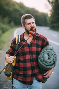 搭便车者走在高速公路旁边。 长胡子的男人背着背包睡觉，吃着棍子上的香肠。 夏天在森林里徒步旅行。