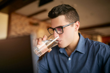 英俊的白种人手里拿着一杯水与笔记本电脑一起工作。戴眼镜的商人在工作时饮用水来进行身体水化。有吸引力的设计师解渴。健康生活方式主题