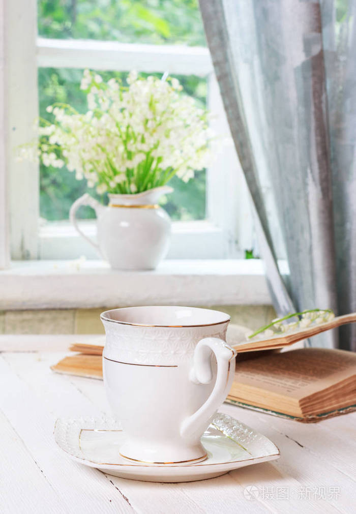 杯茶或咖啡,一本书,放在一张白色的木制古董桌子上,一束山谷的百合花