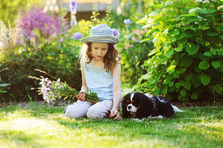 快乐的女孩在夏天的花园里放松，她的猎犬戴着园丁帽，拿着一束花。 背景上盛开的多年生植物。图片