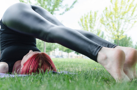 美丽的红头发女人在公园蚂蚁或下面的草地上练习瑜伽。 运动的女人在大自然中做伸展运动。 健康的生活和健身理念。