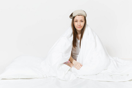 有趣，疲惫的年轻女人坐在床上，用睡眠面罩，床单，枕头包裹在毯子上，孤立在白色的背景上。 美女花时间在房间里。 休息放松好心情的概