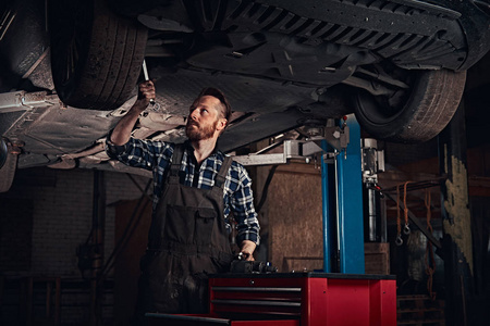 有胡子的汽车技工在制服修理汽车在修理车库的起重车站立下用扳手暂停。