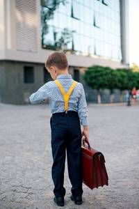 未来商人的概念。 后景男孩穿着深蓝色裤子，婴儿蓝色衬衫和黄色带子站在步行街上，带着布劳恩公文包，看着他的手表城市背景。
