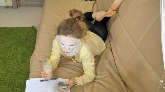 年轻妇女做面膜面具与清洁面具, 点击在沙发上与智能手机在家里