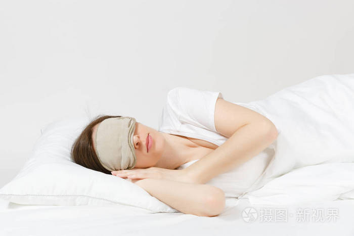 平静的年轻黑发女人躺在床上,白色床单枕毯上的睡眠面罩在白色背景上.