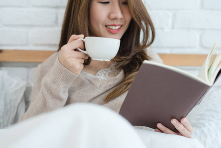 美丽的亚洲女人在卧室的床上享受温暖的咖啡和看书。亚洲女性穿着舒适的毛衣，拿着一本书和一杯咖啡。生活方式亚洲女人在家里的概念。
