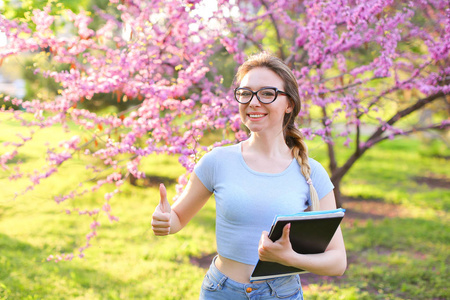 欢快的女学生在盛开的公园里竖起大拇指和保存文件