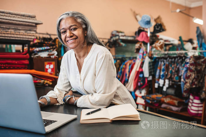 老板在笔记本电脑上工作,并在她被五颜六色的纺织品包围的商店柜台写