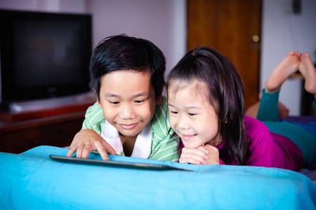 亚洲儿童一起观看数字平板电脑学习。 中国男孩和他的妹妹微笑着俯卧在床上。 关于在家里使用电子学习技术进行教育的概念。