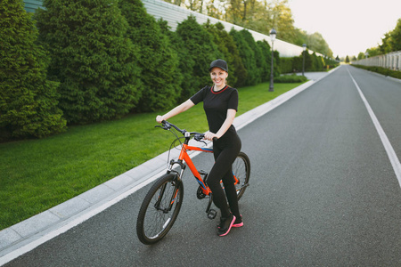 年轻的运动黑发坚强的女人，穿着黑色制服的帽子，在春天或夏天阳光明媚的日子，停止骑黑色自行车，在户外骑橙色元素的黑色自行车。 健身