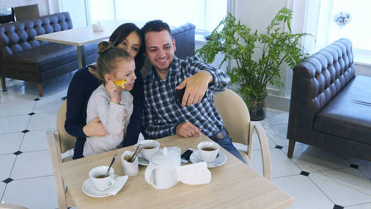 快乐的父亲需要自拍照照片与他的妻子和女儿在他们在咖啡馆或餐厅的下午茶时间