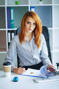生意。 商务女性红头发，坐在大光办公室的桌子旁，手里拿着一个文件夹，写字。 穿着白色衬衫和黑色裙子的女孩。 严肃女人的肖像。