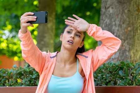 穿着运动服的拉丁女人穿着运动服在一个现代城市公园早上，在做运动后看上去又累又累，在健身和健身中在手机上自拍。