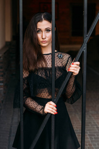 一个年轻漂亮女孩的肖像。 穿着黑色连衣裙的黑发女人站在有黑色棒的笼子附近。 粘在他们身上。 时尚时尚肖像的情感肖像美丽的年轻女子
