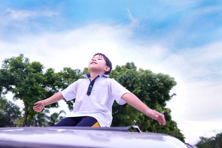 滑稽的亚洲小男孩站在车的天窗上，背景是云树和蓝天。夏天可爱的孩子的微笑和幸福。假期，假期，家庭，旅行概念。