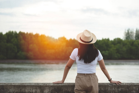 傍晚日落时，戴着帽子的快乐时髦女人站着看河的背面景色。 年轻的亚洲旅行者在自然户外湖享受时光。 生活方式旅行放松和自由的概念。