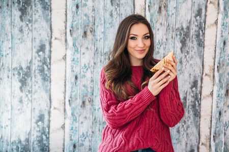 一个可爱的, 开朗的欧洲看起来的女孩穿着时尚的保暖夹克要吃她的三明治满意, 在一个木制的背景下