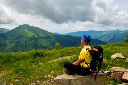 冒险家正在山上的小径上放松，坐在巨大的石头上，欣赏着在低沉的云层下延伸到地平线上的树木山丘的全景。 后景。