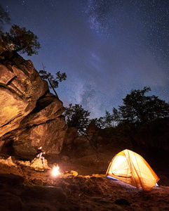 营地之夜，在巨大的陡峭岩层中。 迷人的女人坐在巨石上，在蓝色星空背景下，在旅游帐篷前观看篝火。 旅游体育和野营理念。