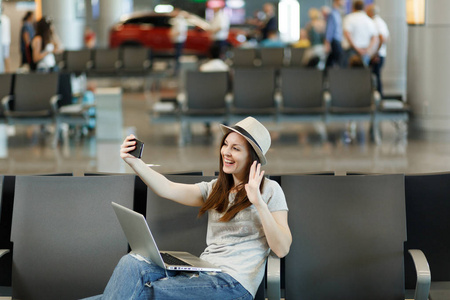 年轻的微笑旅行者旅游妇女在笔记本电脑上工作，在手机上自拍，等待在国际机场大厅。 周末出国旅行的乘客下车。 空中飞行概念