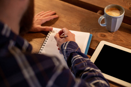 年轻的大胡子商人坐在咖啡馆, 家在桌子上, 写在笔记本上, 靠近谎言的平板电脑与黑屏。人在工作, 学习