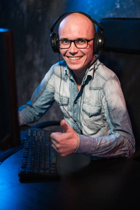一个快乐的年轻玩家的肖像在家里玩电子游戏在电脑上。玩家在耳机是高兴, 并显示竖起大拇指的手势, 因为他赢得了比赛。电脑游戏概念