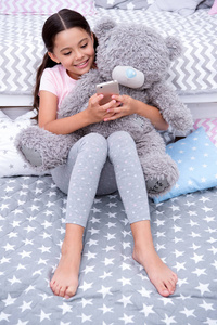 发送消息甜美的梦。女孩坐在床上, 泰迪熊在她的卧室。孩子准备上床睡觉。女孩长头发可爱的睡衣放松毛绒泰迪熊玩具。儿童智能手机发送消
