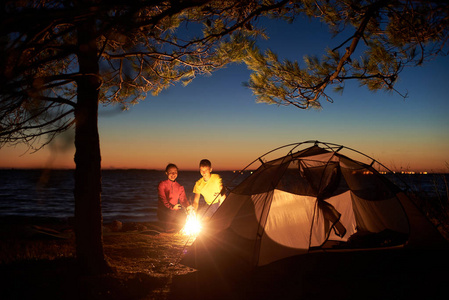 晚上在海上露营。 树下的旅游帐篷和年轻的夫妇在篝火对夜空和橙色的地平线上。 旅游幸福关系和积极的生活方式观念。