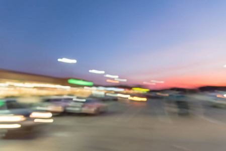 抽象模糊的现代商业地带在欧文德克萨斯美国日落。 购物中心一排汽车在户外无盖停车场，开着零售商店的灯杆，背景是