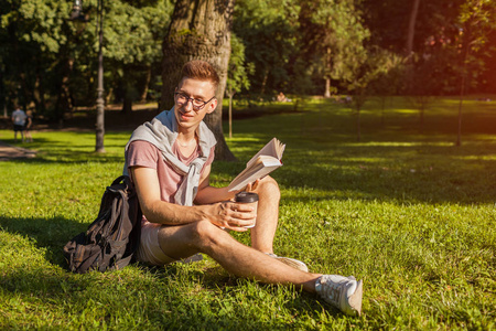 英俊的大学生在校园公园看书和喝咖啡。 快乐的家伙学生在春天坐在草地上户外学习。 教育概念