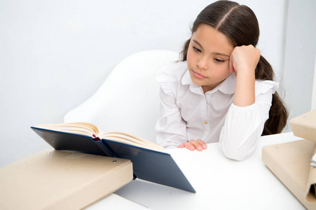 读无聊的书。女孩子看书时坐在桌子白色背景。学生学习和阅读的书。孩子女孩学校制服无聊冷漠的面孔哀伤地读乏味文学