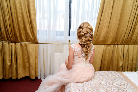 迷人的新娘穿着婚纱，美丽的发型坐在床上，在卧室的窗户。 新娘等待新郎在室内后景