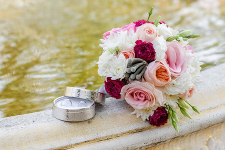 关闭美丽的新鲜婚礼花束粉红色和白色玫瑰和两个金色结婚戒指在盒子上的石头喷泉自由空间。 婚礼细节户外与复印空间