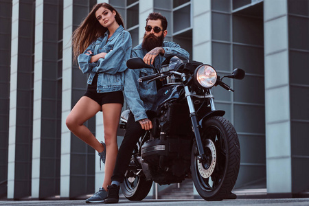 迷人的时髦夫妇留着胡子，穿着太阳镜和牛仔裤夹克，坐在复古摩托车上，他的年轻女孩站在摩天大楼旁边摆姿势。