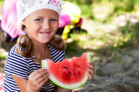 夏天温暖的一天，一个小可爱的小女孩在野餐时吃了一大块西瓜，背景是一片海滩和绿草