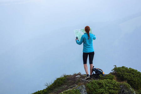 在山顶上，带着地图和指南针的旅游女孩正在寻找梦想的道路。雾中的地平线。夏天的风景。极限运动。