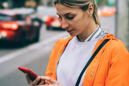 关闭年轻女性使用手机数据互联网连接，同时站在城市街道。 穿橙色夹克的白种人时髦女孩在智能手机应用程序上打电话给出租车