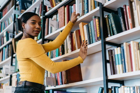 美国黑人女青年穿着休闲装，一边看着相机，一边从公共图书馆的书架上拿着文学书籍。深色皮肤的学生选择学习课本