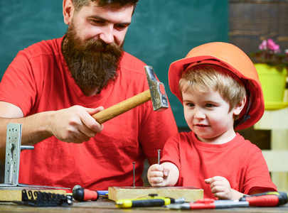 男孩, 孩子忙着保护头盔学习用锤子和爸爸在一起。父亲用胡子教小儿子用工具, 锤击, 黑板上的背景。杂工概念