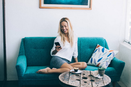 积极的女性博客作者通过应用程序在智能手机上输入短信，同时在舒适的家沙发上休息，快乐的时髦女孩通过移动应用程序在社交网络上观看有趣
