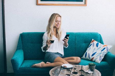 快乐的金发碧眼的时髦女孩享受咖啡时间，通过手机和朋友在室内传递信息，微笑的女人躺在舒适的家沙发上，一边在网上阅读新闻