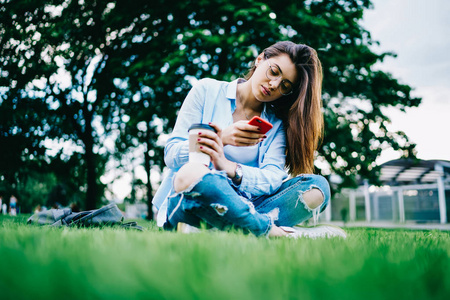 有思想的年轻女性，长着深色头发，通过智能手机查看收入信息通知，坐在公园的草地上，女学生在户外喝咖啡，通过手机聊天，在校园里聊天。