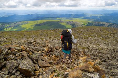 一个女游客头上戴着一个大背包，手里拿着一根木棍，从山上下到岩石上，面前是一个绿色的山谷，有着蓝天和白云