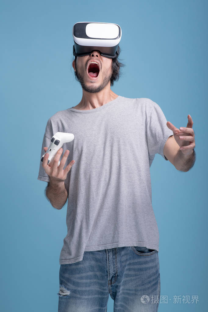 年轻的胡须男在蓝色背景下玩虚拟现实游戏令人震惊的亚洲年轻人戴着vr