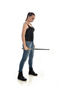 穿黑色单身牛仔裤的黑发女孩的全长肖像。 站着摆姿势拿着剑。 孤立在白色工作室背景上。